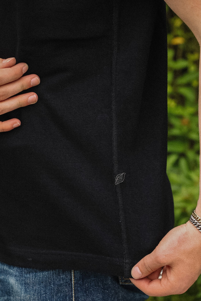 Close-up of a person wearing a black short-sleeve knit top with blue jeans, showing the side seam and a small diamond-shaped logo patch near the hem.