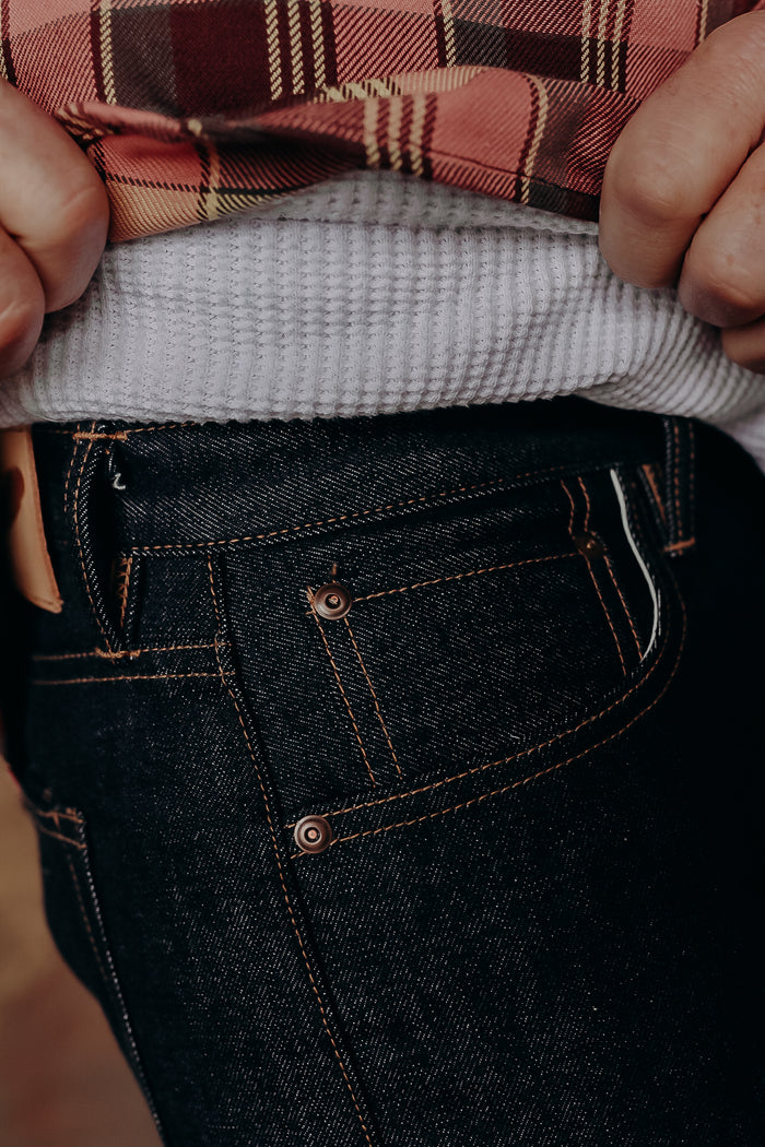 Close-up of person wearing dark denim jeans and lifting plaid shirt to show white textured undershirt