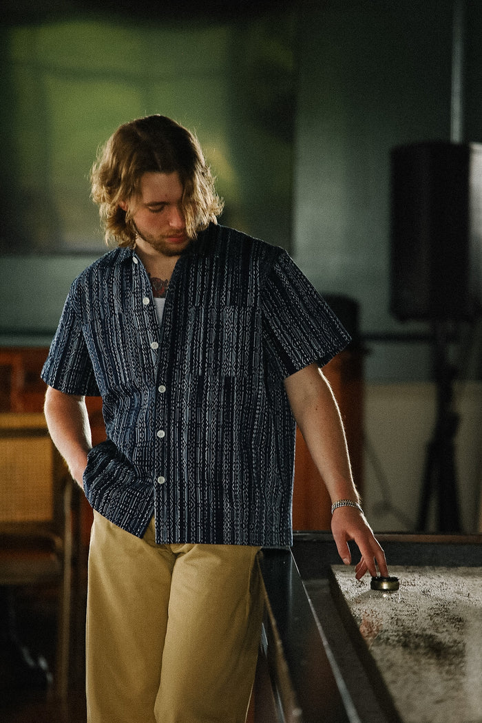 Man in a dark patterned shirt and beige pants standing near a pool table.