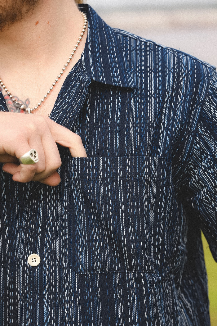 Close-up of a person wearing a dark blue patterned shirt with a necklace and ring.