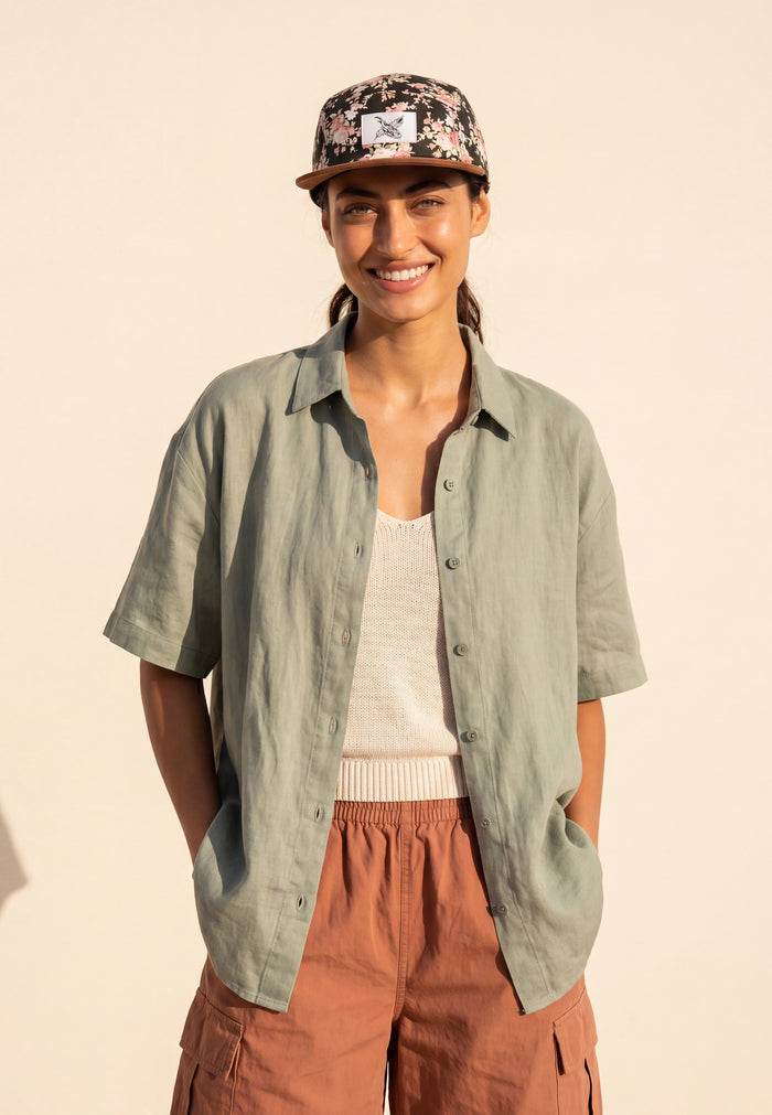 Woman wearing a green shirt, brown pants, and a patterned cap against a beige background