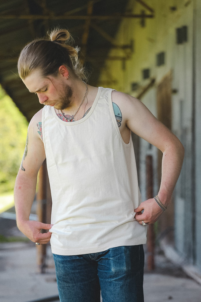 A man with a bun and beard wearing an off-white tank top and blue jeans, standing in front of a rustic building.