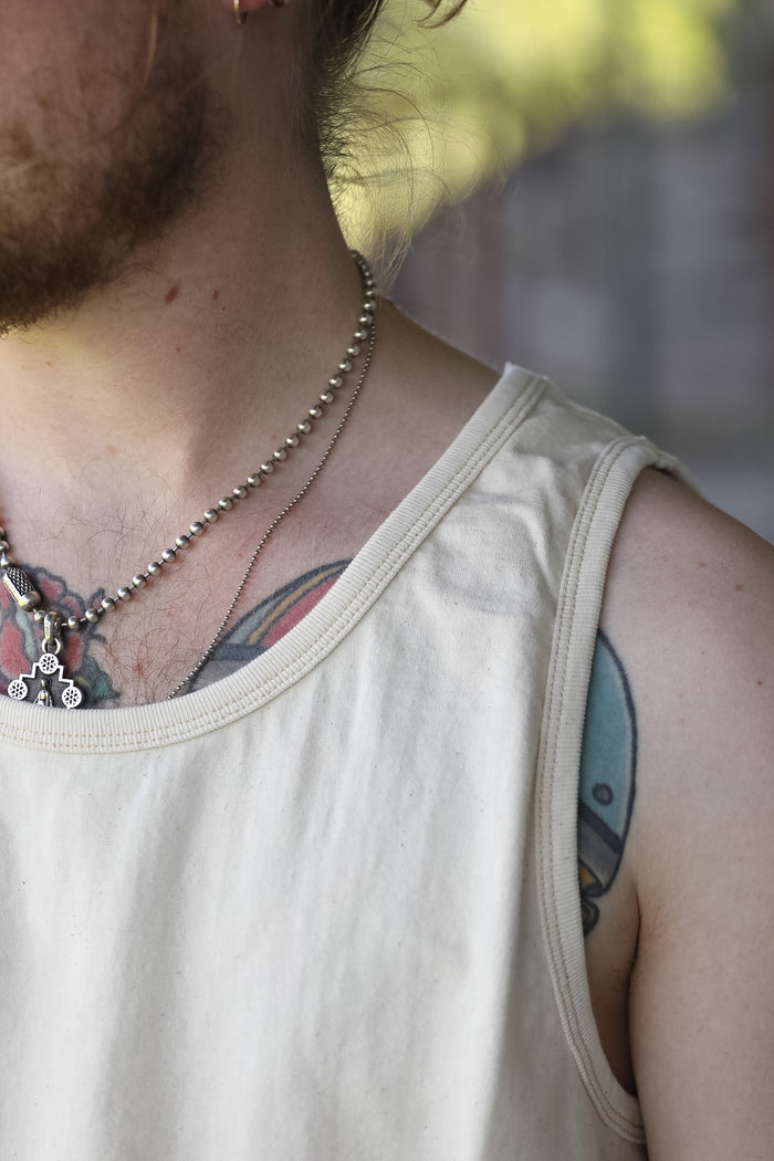 A close-up shot of a man's neck and upper chest wearing a silver ball chain necklace with a small pendant, layered over a light beige tank top.