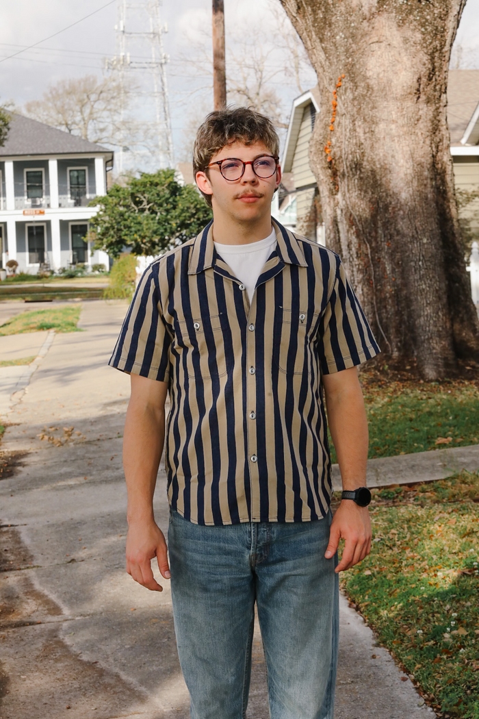 Person wearing a striped shirt and jeans standing on a sidewalk with a tree and house in the background.