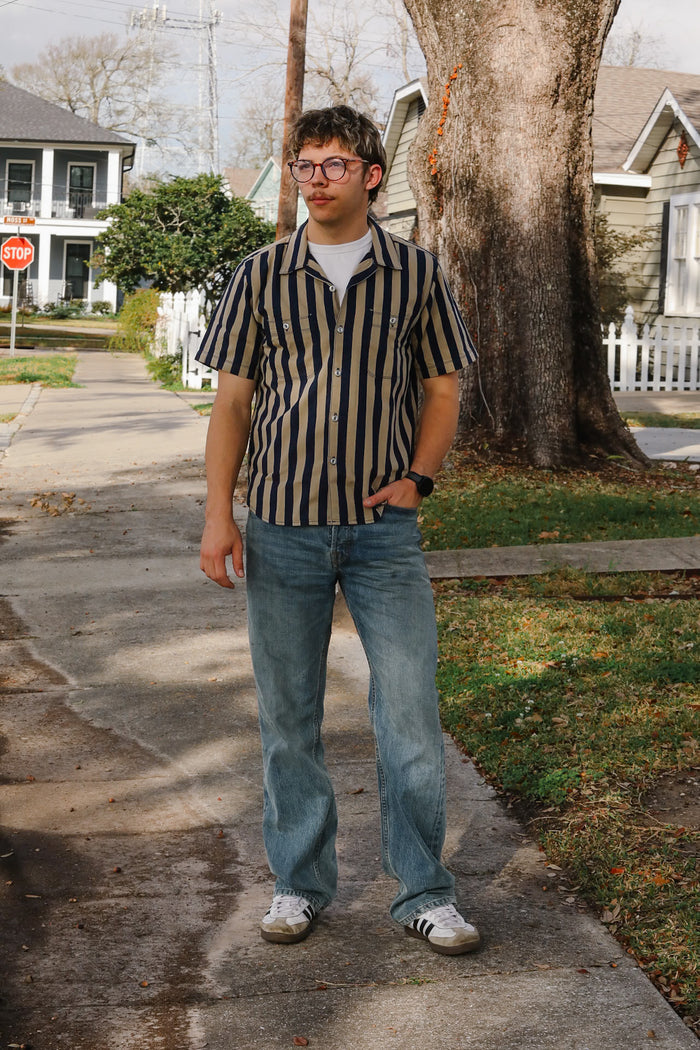 Person wearing a striped shirt and jeans standing on a sidewalk with houses and trees in the background.