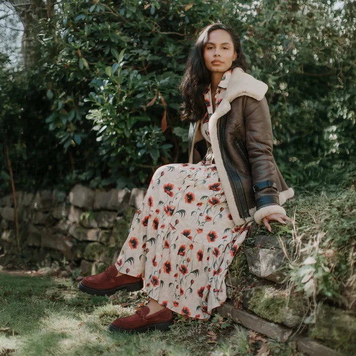 Woman in a floral dress and brown coat sitting outdoors among greenery