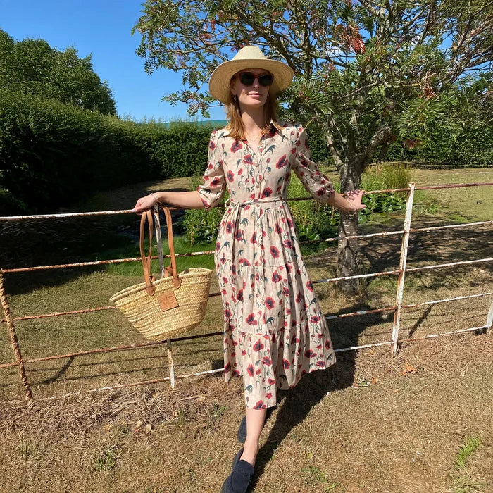 Woman in a floral dress and sun hat standing by a fence in a garden.