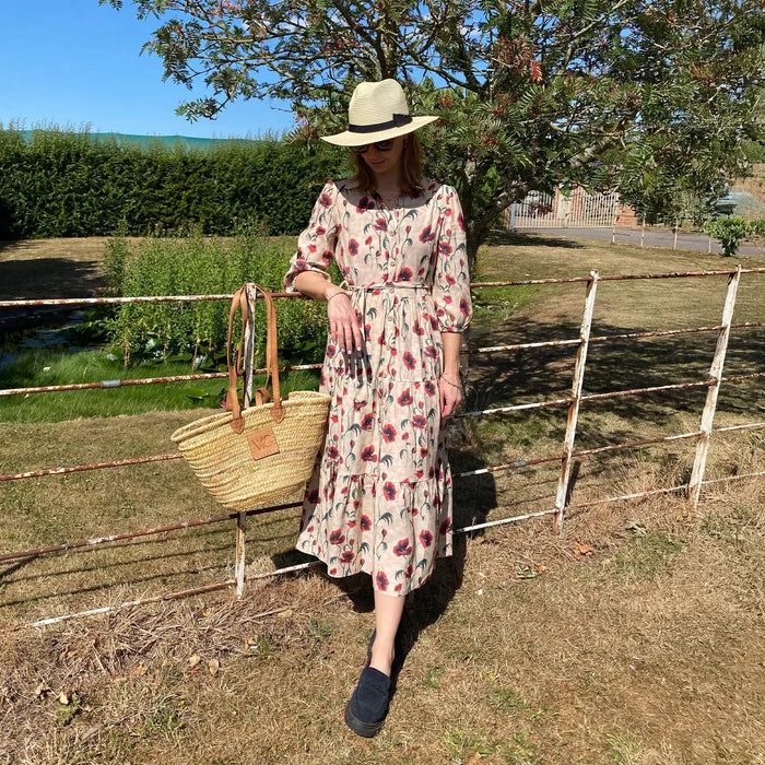 Woman in a floral dress and hat standing by a rustic fence outdoors.