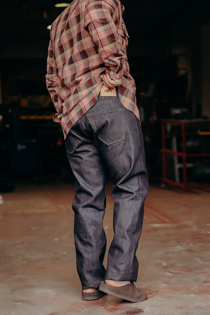 Person wearing dark raw denim jeans and a red plaid shirt in a workshop setting
