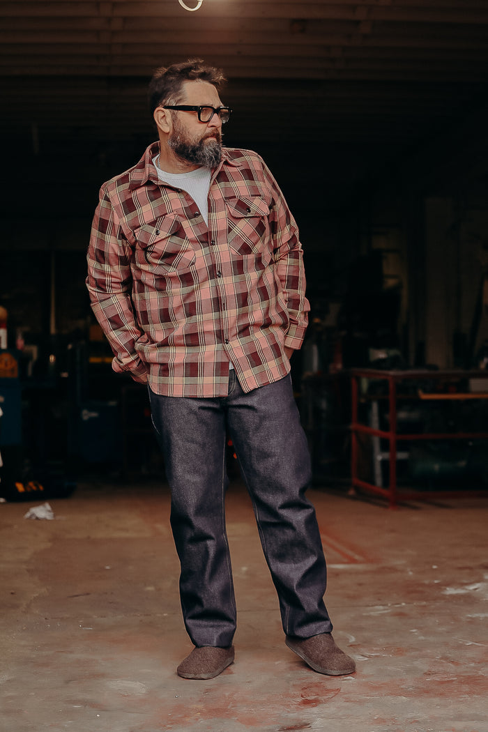Man wearing brown plaid shirt, raw denim jeans, and brown slippers in workshop setting